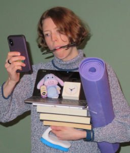 Middle-aged person with a pen in the mouth holding a phone in one hand and an exercise mat, laptop, clock, books, notebook, scrub brush, and an Eeyore plushie in the other.