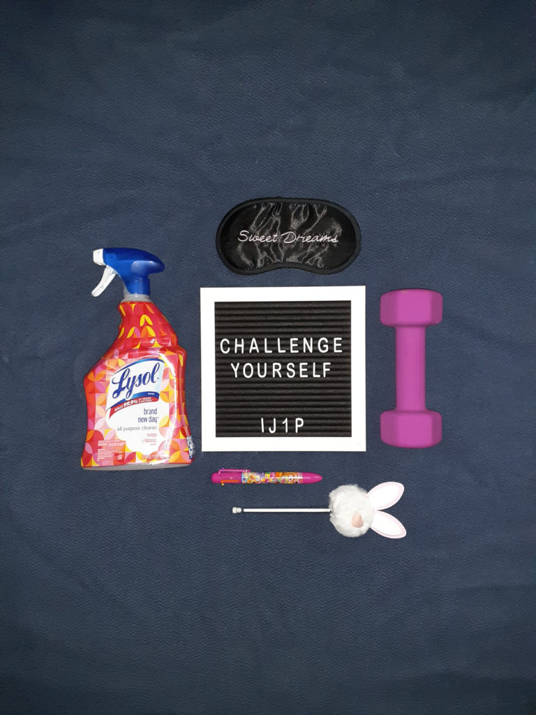 A board reading "challenge yourself" surrounded by cleaning supplies, a weight, a sleep mask, and two pens.