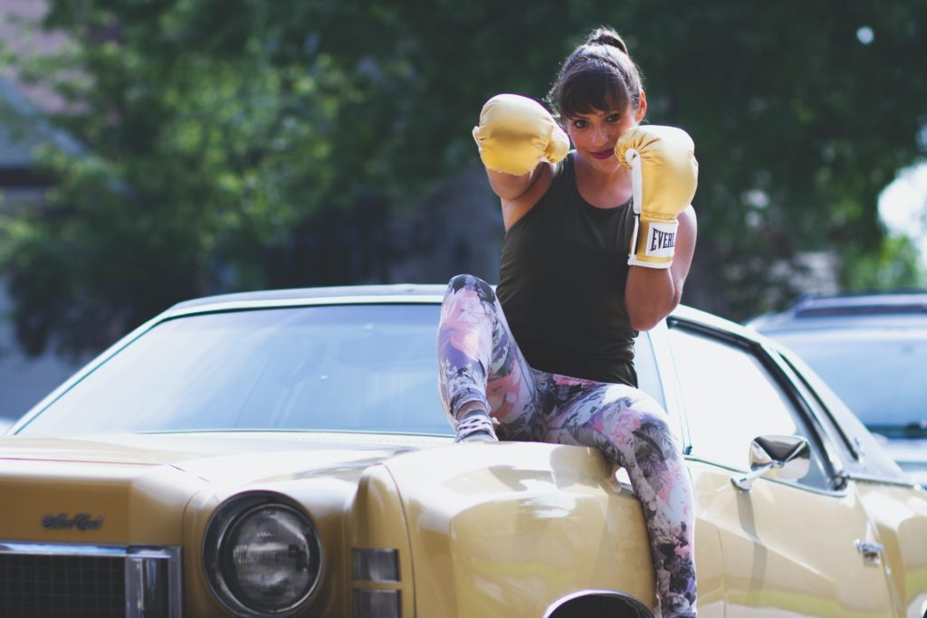 Woman sits on hood of car, wearing boxing gloves